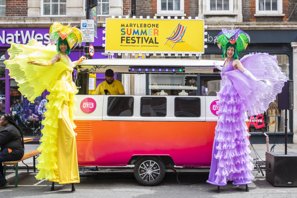 Female stilt walkers dressed in bright purple and bright yellow frills stand smiling on either side of a street food truck at Marylebone Summer Festival, produced by We Are Placemaking for the Howard de Walden Estate