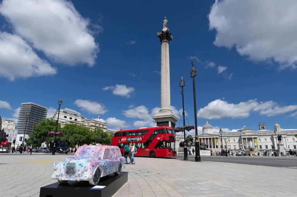 Colourful black cab sculture positioned in front of Trafalgar Square, as another London icon, the red bus, drives past