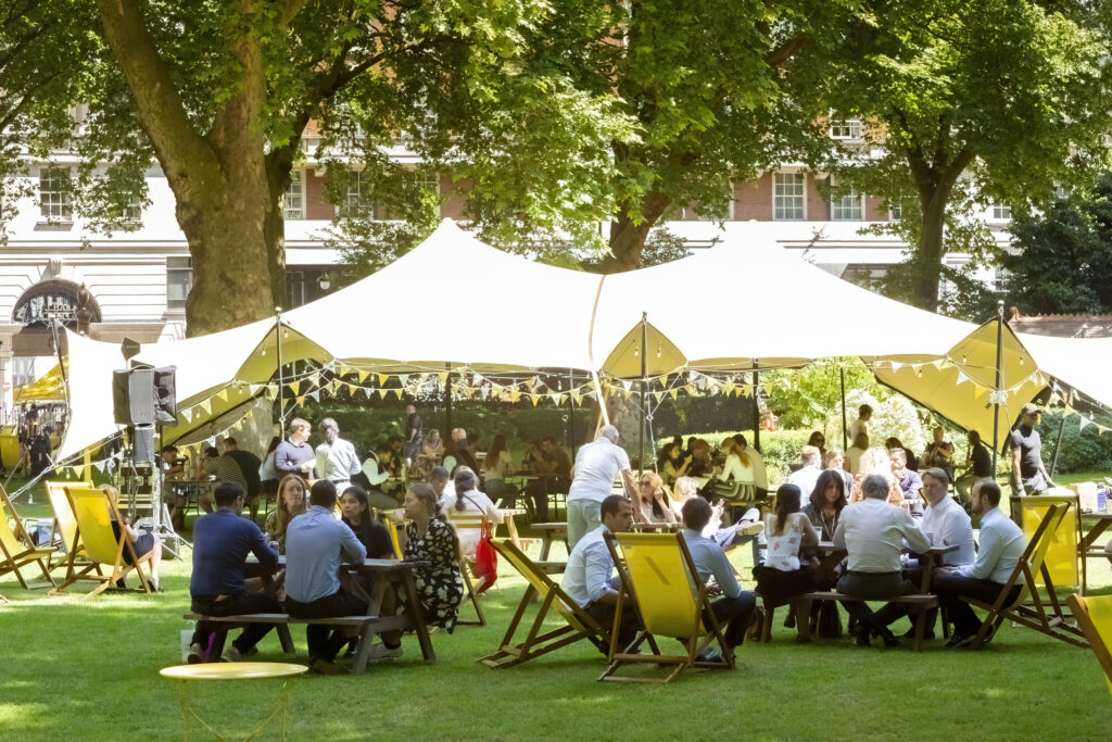 Visitors gather on benches and deck chairs near a yellow marquee dressed in bunting set up in Portman Square by We Are Placemaking for the Baker Street Summer Series