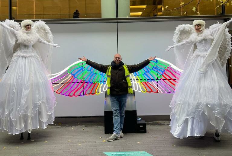 We Are Placemaking Event Producer Will posing with two angels on stilts and in front of a light-up angel wings installation